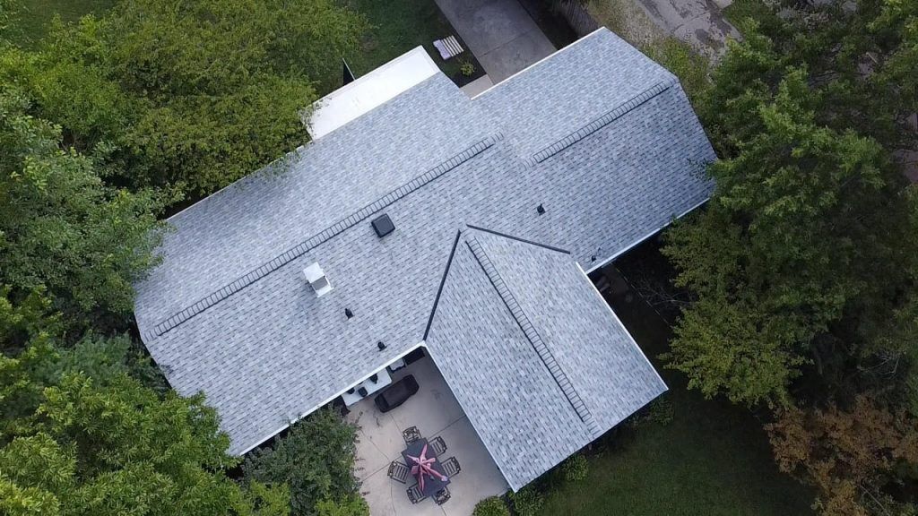 Aerial view of a large house with a grey shingle roof, surrounded by greenery, with a carport and a red object on the ground.